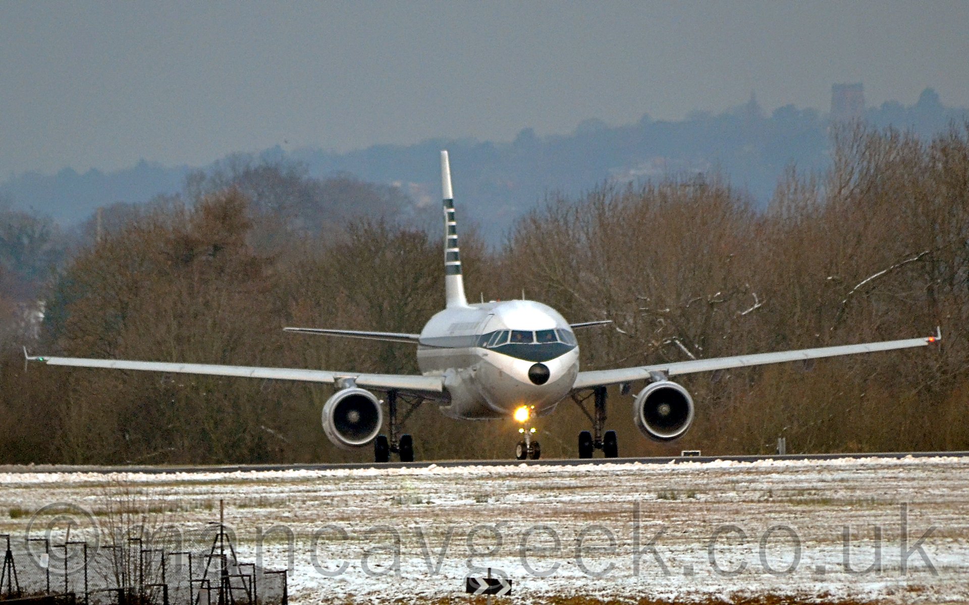 Almost head-on shot of a white and dark green, twin engined jhet airliner, taxiing from left to right, pointing just off to ther right of the camera, the landing light on the nosewheel flaring down the lens. A light dusting of snow covers grass in the foreground, with trees in the background, under a grey sky.