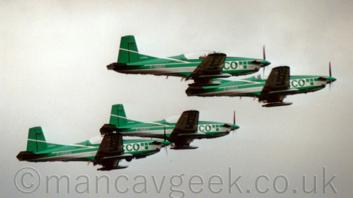 Side view of 4 green and white, single engined trainer aircraft flying from left to right in formation, against a backdrop of a flat grey sky.