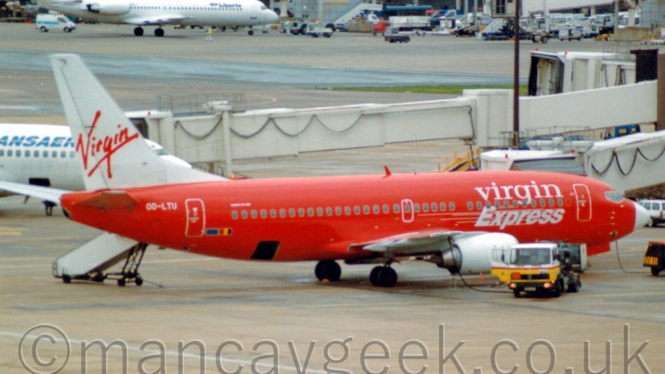 Side view of a bright red, twin engined jet airliner with a white tail parked facing to the right, with air bridges and another couple of planes in the background.