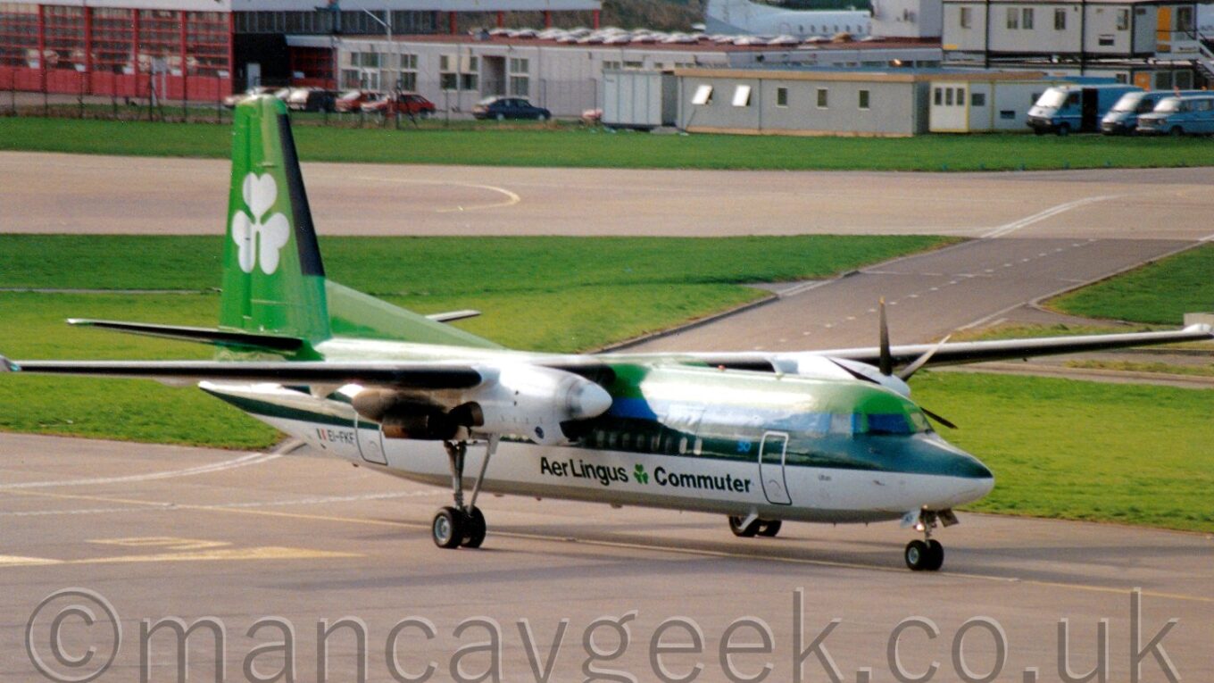 Side view of a green and white, high-winged, twin propellor-engined airliner taxiing from left to right, with low, grey buildings in the background.