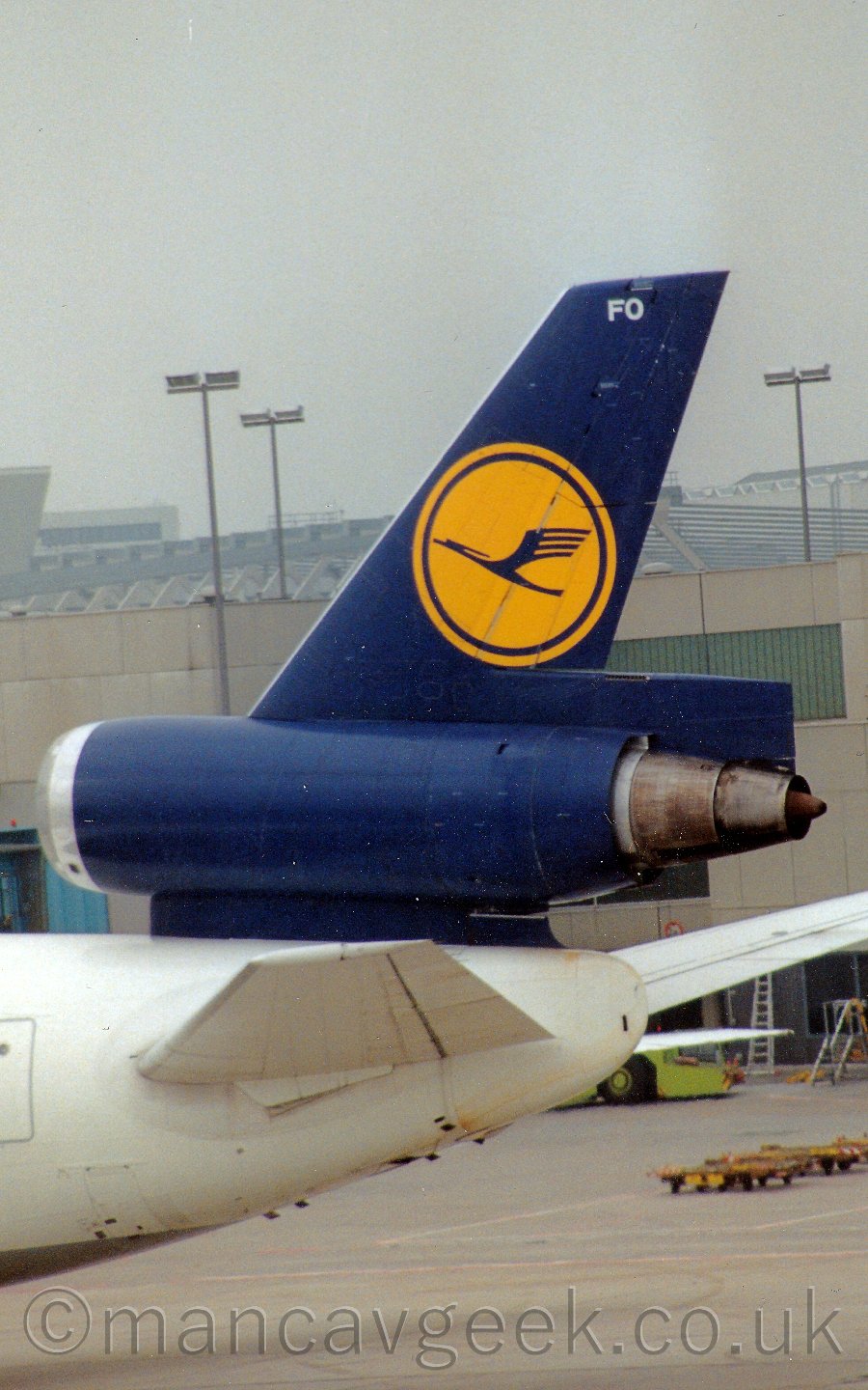 Closeup of the blue and yellow tail and centre engine of a white, 3 engined jet airliner parked facing to the left, with airport buildings in the background, under a dismal grey sky.