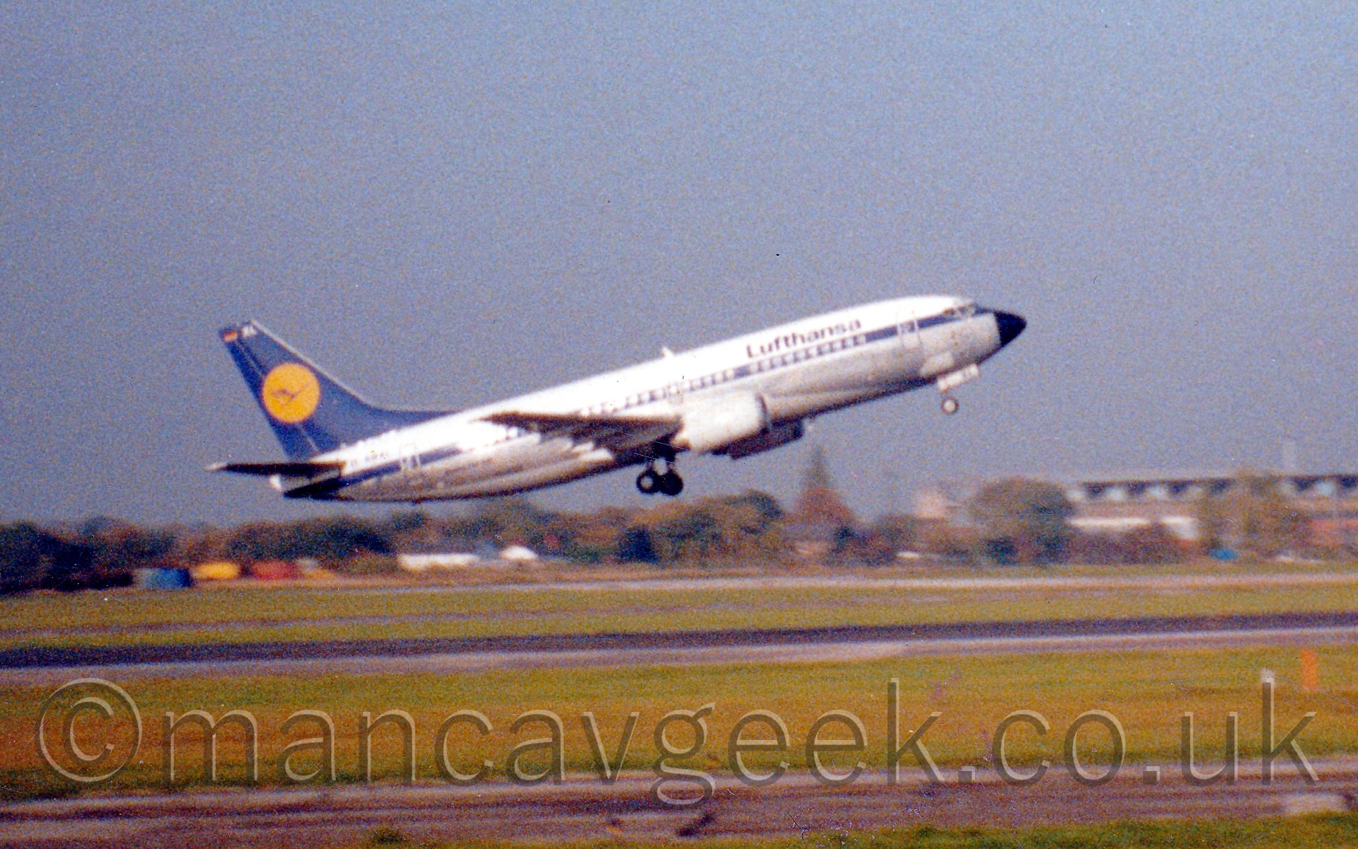 Rather grainy side view of a white, silver, and blue, twin engined jet airliner flying from left to right just feet above a runway, it's nose raised around 30 degrees, suggesting it has just taken off, with green grass and grey taxiways in the background stretching off to rows of trees in the distance, under a hazy grey-blue sky.