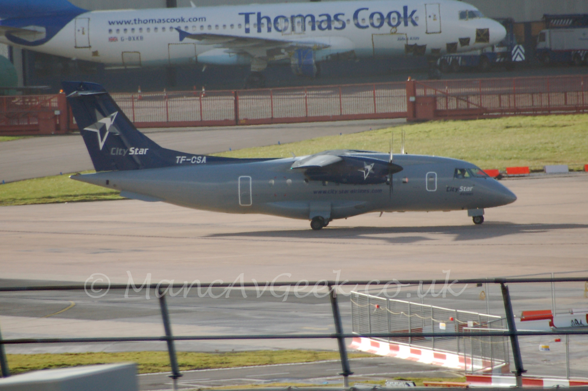 Side view of a dark grey, high-winged, twin propellor-engined airliner with dark blue tail and engine pods, with a white and blue plane behind a red metal fence in the background.