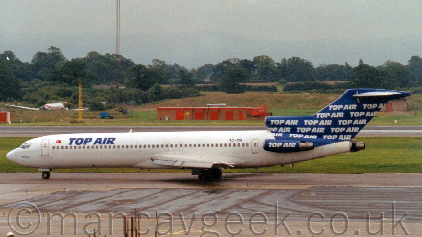 Side view of a white, 3 engined airliner with the engines mounted on the rear fuselage, and a blue and white tail, taxiing from right to left, with tree-lined fields in the backgroud, under a grey sky.