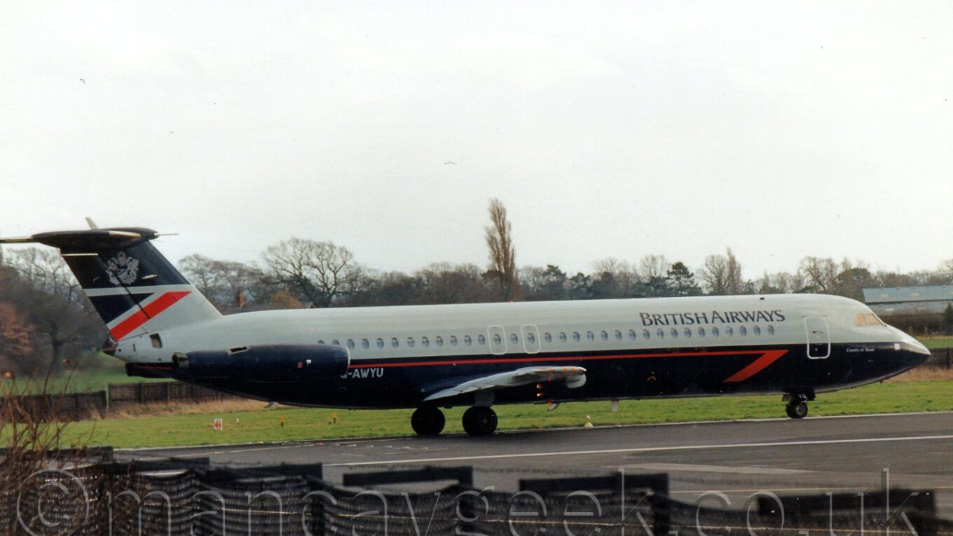 Side view of a dark blue and grey, twin engined jet airliner with the engines mounted on the rear fuselage, taxiing from left to right on a runway, with a black chain link fence in the foreground, and grass and trees in the background, under a grey sky.