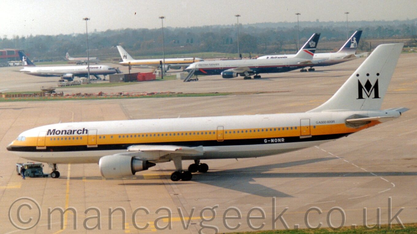 Side view of a white and yellow, twin engined jet airliner facing to the left but being pushed backwards to the right by a smlal blue tug attached to the nose wheel, with more planes in the backgrounds, under a hazy grey sky.