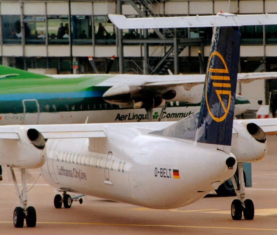 Side view of a white, high-winged, twin propellor-engined airliner taxiing from right to left with a green and white airliner in the background next to a heavily glazed building, under a bright sky.