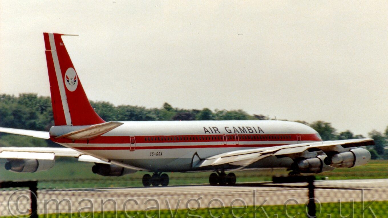Side view of a white and red, 4 engined jet airliner moving from left to right on a runway, with trees peeking over the top of the fuselage, under a bright but hazy sky.