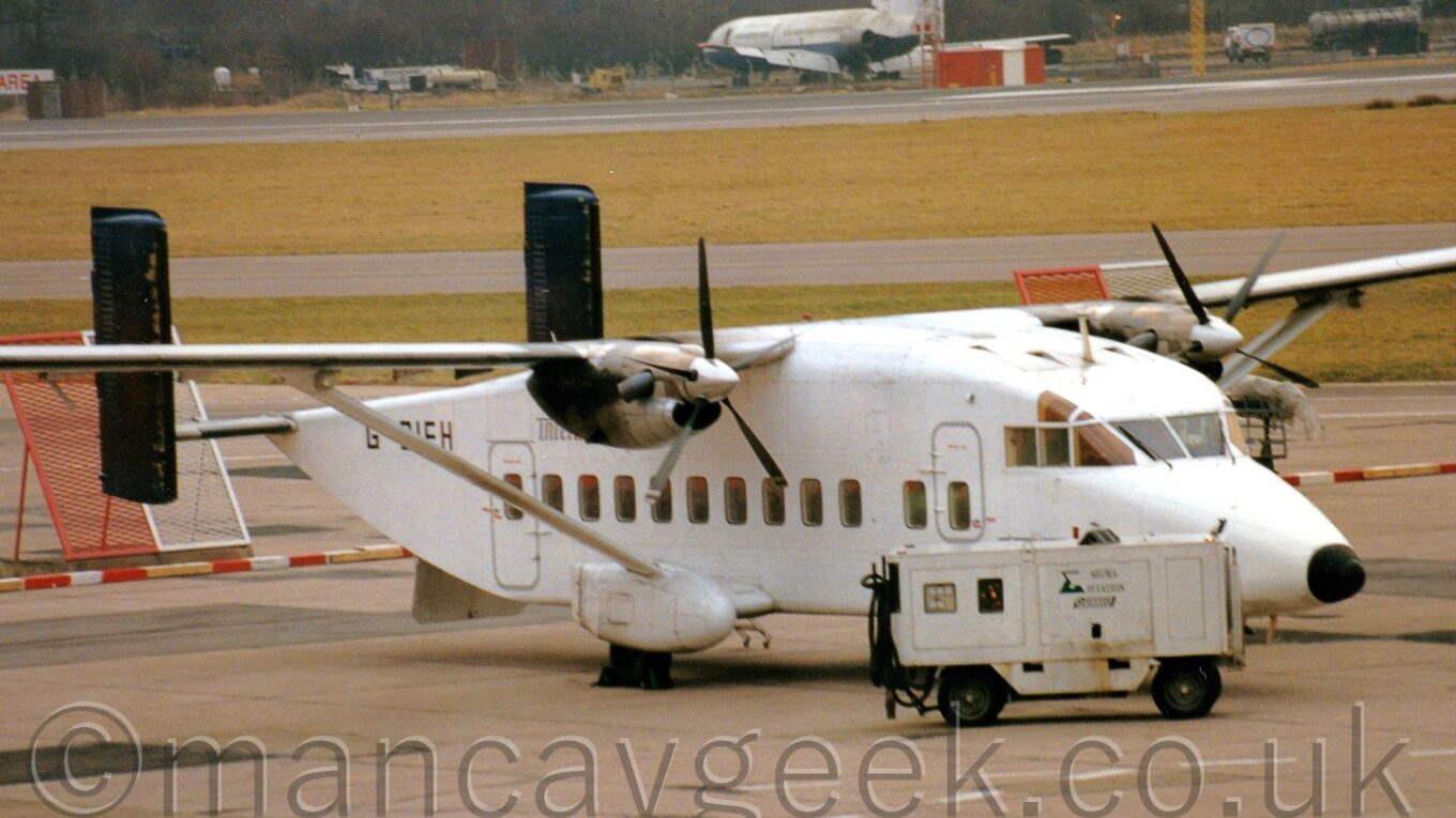 Side view of a white, high-winged, twin-tailed, twin propellor-engined airliner parked facing to the right, with a portable power unit parked uin front of the plane's nose, and a derelict white and dark blue airliner in the far background.