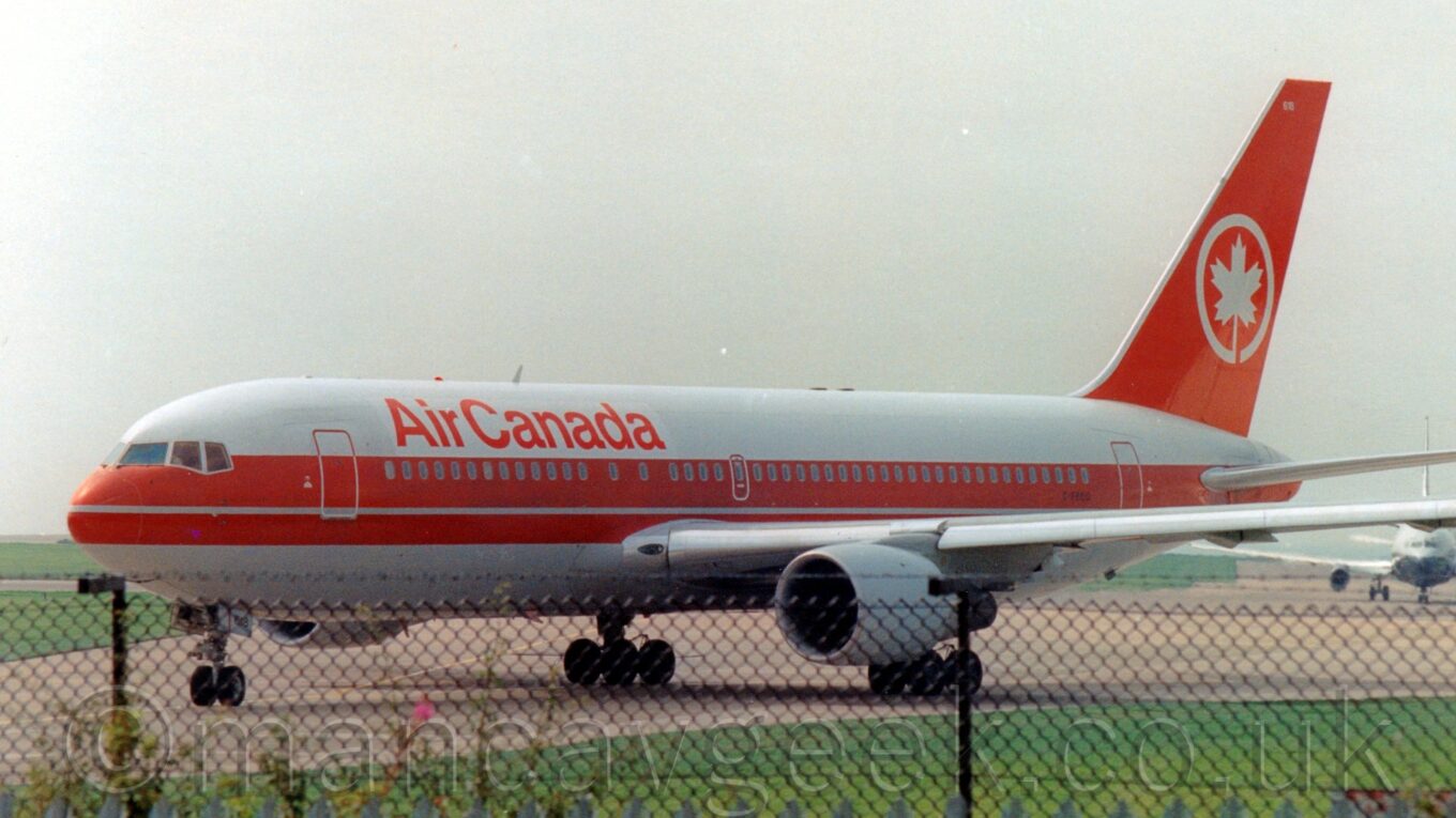 Side view of a white, twin engined jet airliner with a pair of red stripes running along the fuselage and a red tail, taxiing from right to left, turning slightly away from the camera, with a black chainlink fence and grass in the foreground, and hazy grey sky filling the rest of the frame.