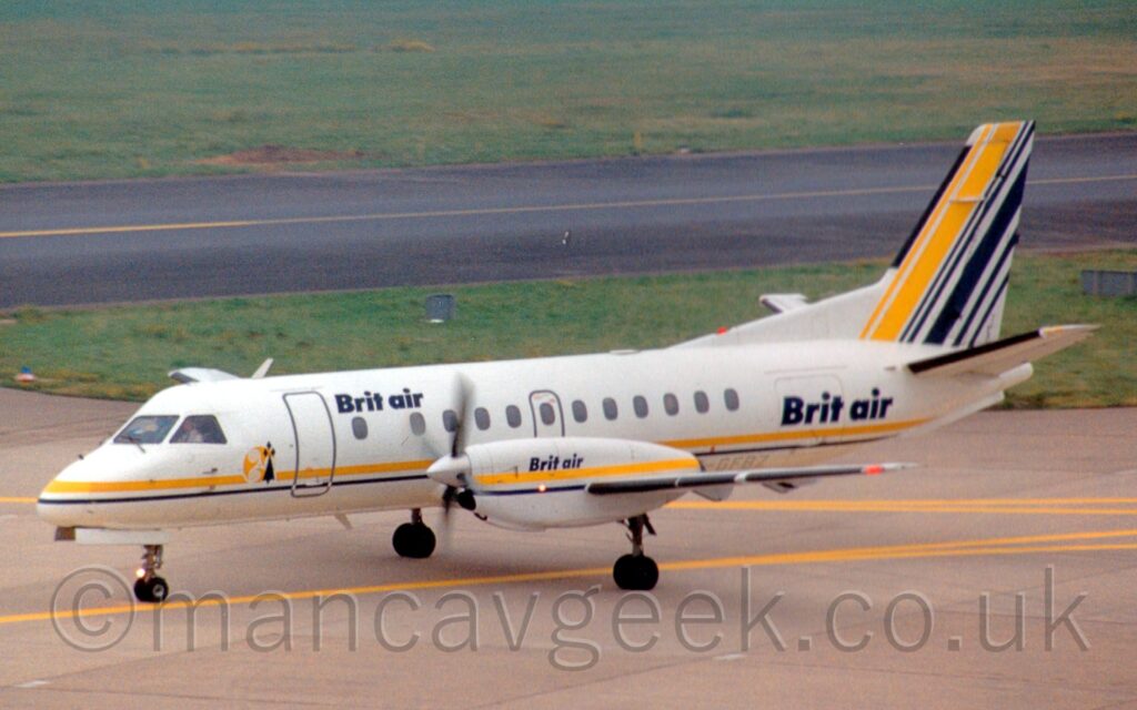 Side view of a twin propellor-engined airliner taxiing from right to left, following a yellow line marked on a grey taxiway. The plane is white, with a yellow and black stripe running along the lower body, just above the wing root, as well as along the engine pods on the wings. There are black "Brit Air" titles on the upper forward fuselage just aft of the main door, on the rear fuselage , covering the rear baggage door, and on the sides of the engine pod. There are a series of yellow and black diagonal stripes on the tail. Green grass fills the background, with a black taxiway cutting through from side to side.