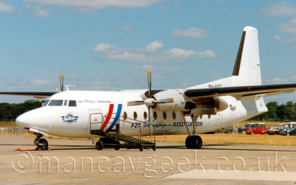 Side view of a high-winged, twin propellor-engined airliner parked facing to the left on grey concrete apron, with a rather strange looking green set of airstairs parked by it's forward door. The plane is mostly white, with diagonal red, white, and blue bands on the sides of the forward fuselage, representing the Dutch flag, and black titles "F27 Friendship Association" on the lower body, more or less centred under the wings, with a smaller variant of those titles also on the upper forward fuselage, just aft of the cockpit. A logo on the nose, under the cockpit windows, shows an image of one of these planes flying around a stylised wire-frame globe, with 3 blue stripes in the background, thick and the top and thin at the bottom. The registration "PH-KFG" is on a fillet at the front of the tail, above the rear fuselage, partially obscured by the wing. Grey concrete apron fills the foreground, with a large grassed area being partially used as a car park in the background on the right, and trees in the distance under blue sky with occasional fluffs of white cloud.