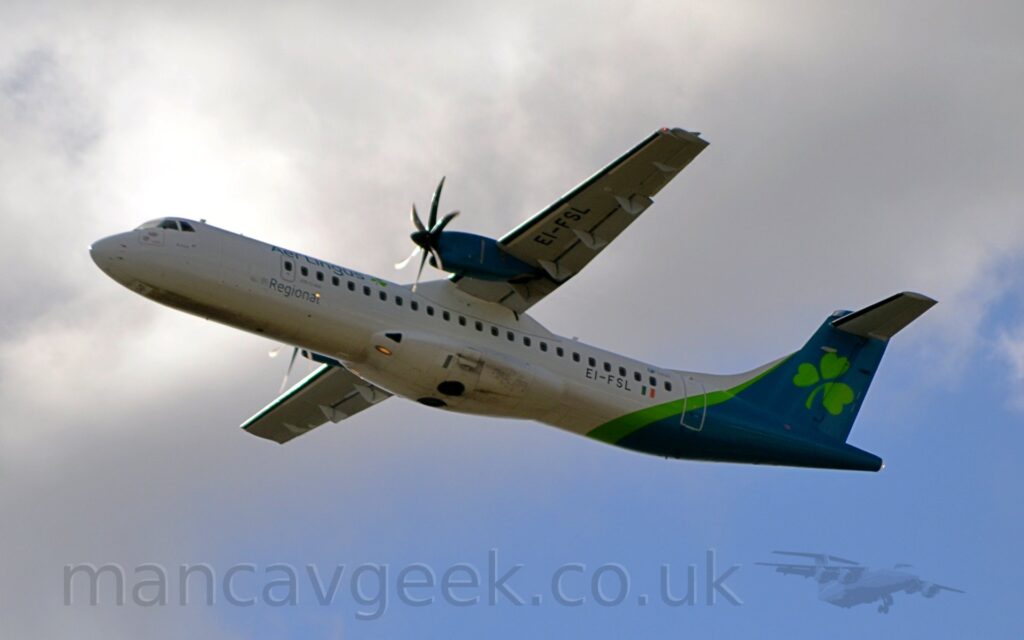 Low side view of a high-winged, twin propellor-engined airliner flying from right to left, with it's undercarriage already tucked away, flaps extended from the rear of the wings, and the nose raised sharply, suggesting it has just taken off. The plane is mostly white, with green "Aer Lingus" titles on the upper forward fuselage, with grey "Regional" titles below. The rear fuselage and tail is a dark blue-green, with a vivid diagonal green stripe in front, and a large green shamrock on the tail. The black registration "EI-FSL" is on the lower rear fuselage, and under the left wing. Grey and white clouds fill most of the background, with a bright spot directly behind the cockpit, and clear blue sky in the bottom right corner.