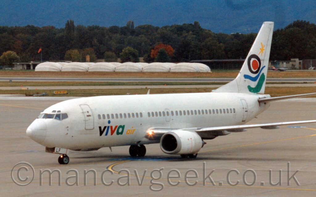 Side view of a twin engined jet airliner taxiing from right to left, following a yellow stripe marked on a large expanse of grey concrete apron that fills the foreground and part of the background. The plane is almost entirely white, with multi-coloured "Viva Air" titles in black, blue, red, green, and yellow, on the lower forward fuselage. Green and blue swirls under black and red circles and a yellow asterisk adorn the white tail. Extensive strips of green grass separate the apron from a taxiway and runway in the background, with white domed tent-style structures on a large grass patch in front of trees on the left of the background. A tree-covered hill rises in the far distance to fill the rest of the frame.