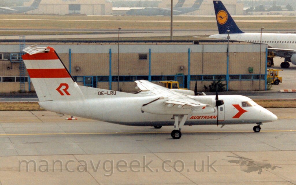 Side view of a high-winged, twin propellor-engined airliner taxiing from left to right. The plane is mostly white, with a grey belly, and red "Austrian" titles on the lower forward fuselage, with a red arrowhead just below and aft of the cockpit. There are a pair of thick red stripes at the top of the white tail, with a stylised red upper-case letter "R" at the base. A pale brown building stretches across the background, with a white jet airliner with a blue and yellow tail just taxiing out of frame at the top right. Large, green, 4 engined military cargo planes are clustered around a cream coloured hangar in the distance, slowly vanishing in to haze.