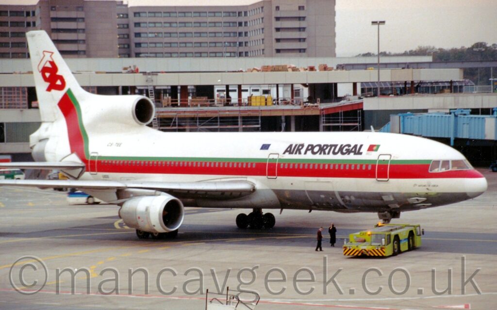 Side view of a white, 3 engined jet airliner facing to the right but being pushed back to the left by a lime green truck attached to the nose wheel. The plane is mostly white, with a bare metal belly, and a thick red and thin green stripe running along the body, covering the passenger cabin windows, before sweeping up into the tail, where it ends with the red and white letters "TAP". There are black "Air Portugal" titles on the upper forward fuselage. A building appears to be under construction in the background, with a taller grey building beyond that.