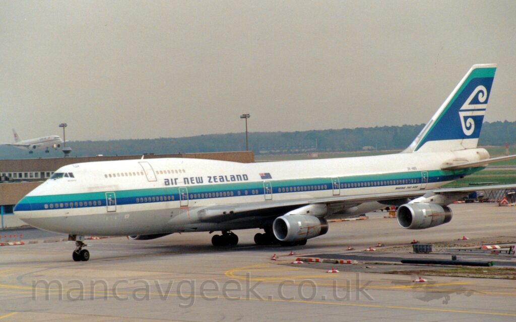 Side view of a very large, 4 engined jet airliner taxiing from right to left. The plane is mostly white, with an aquamarine and dark blue stripe running along the body, and dark blue "Air New Zealand" titles on the upper forward fuselage, next to a New Zealand flag. The dark blue registration "ZK-NBS" is on the upper rear fuselage above the stripe, with text "Boeing 747-400" below. The tail is mostly dark blue with an aquamarine stripe on top and at the front, with a stylised white arrowhead representing wind and waves coming from the rear. Grey concrete apron fills the foreground, with red and white marker blocks delineating an unusable section on the right. A brown building stretches across the frame from the left, with a twin engined white airliner flying at a low altitude behind it, on approach to one of the runways in the distance. Trees on the skyline meet grim grey sky.