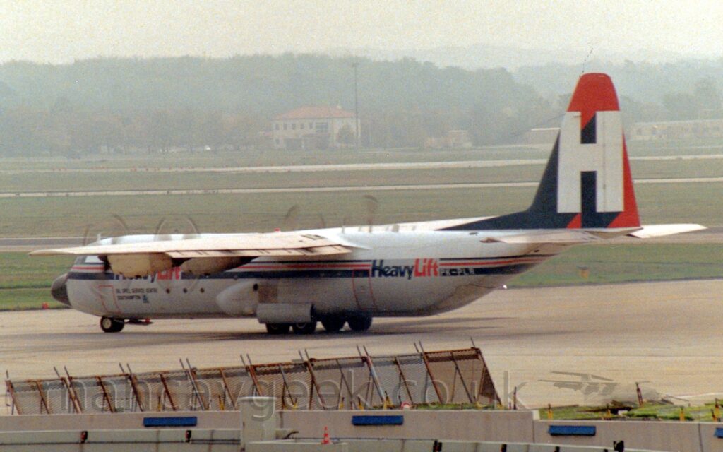 Side view of a high-winged, 4 propellor-engined cargo plane taxiing from right to left and away from the camera. The plane is white on top and has a grey lower half, with a thin red stripe outlined by blue stripes running along the body, separating the 2 colours. There are large, blue and red "HeavyLift" titles overlaid on the stripe on both the forward and rear fuselage, with the white registration "PK-PLS" on the bottom blue stripe right at the rear of the fuselage. The tail is red, with a large white upper-case letter "H", with a massive dark blue drop shadow that fills the front of the tail. Black spinning propellors can be seen making arcs on the front of each engine from over the top of the wing. A metal frame and a grey concrete wall are in the foreground, with grass in the background stretching out to runways in the distance, with buildings beyond that, and trees, slowly being swallowed up in the hazy grey sky.
