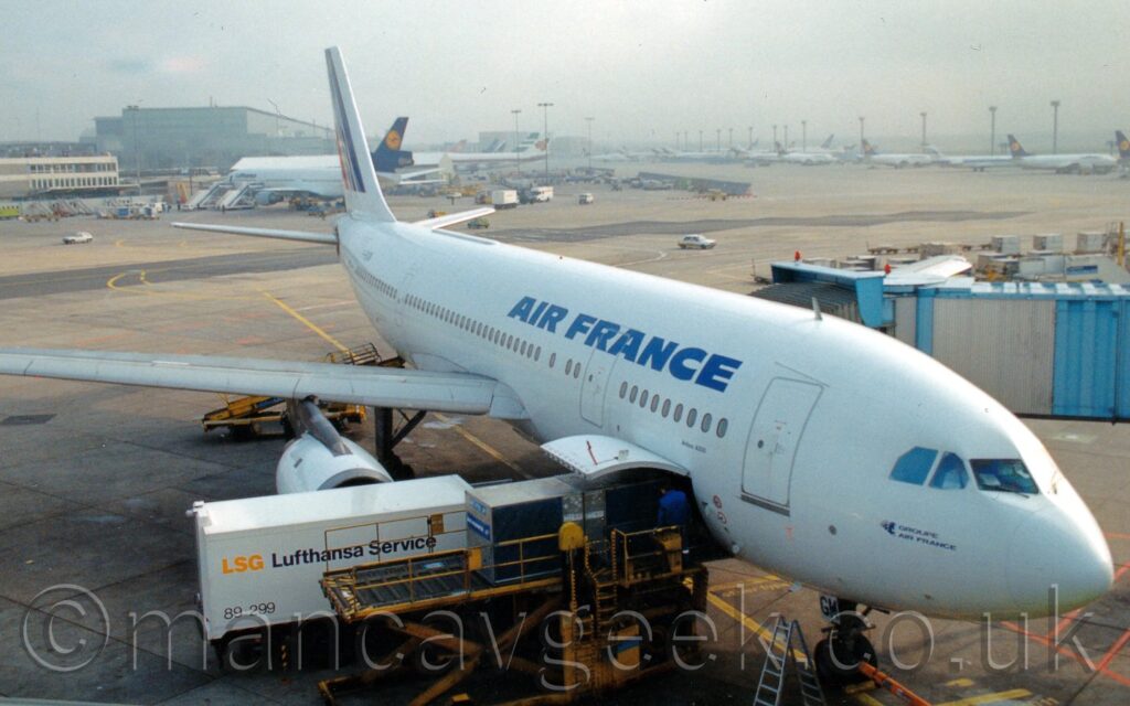Front side view of a twin engined jet airliner parked facing to the right on it's gate almost directly below the camera. The plane is almost entirely white, with blue "Air France" titles on the upper forward fuselage, and a series of diagonal blue stripes on the tail. The forward cargo hold door in the lower forward fuselage is open, with a loading belt raised to it, with a couple of metal containers rolling into the plane's belly, overseen by a person wearing blue overalls. A white truck with black "Lufthansa Service" text on the side is parked between the loading belt and the engine. A pale blue airbridge is attached to the forward door on the other side of the plane, reaching out of frame on the right. A large expanse of apron fills the background, with rows of planes parked by tall lighting towers on the right of the frame, and more planes parked facing a black terminal building on the left, under bright but hazy sky.