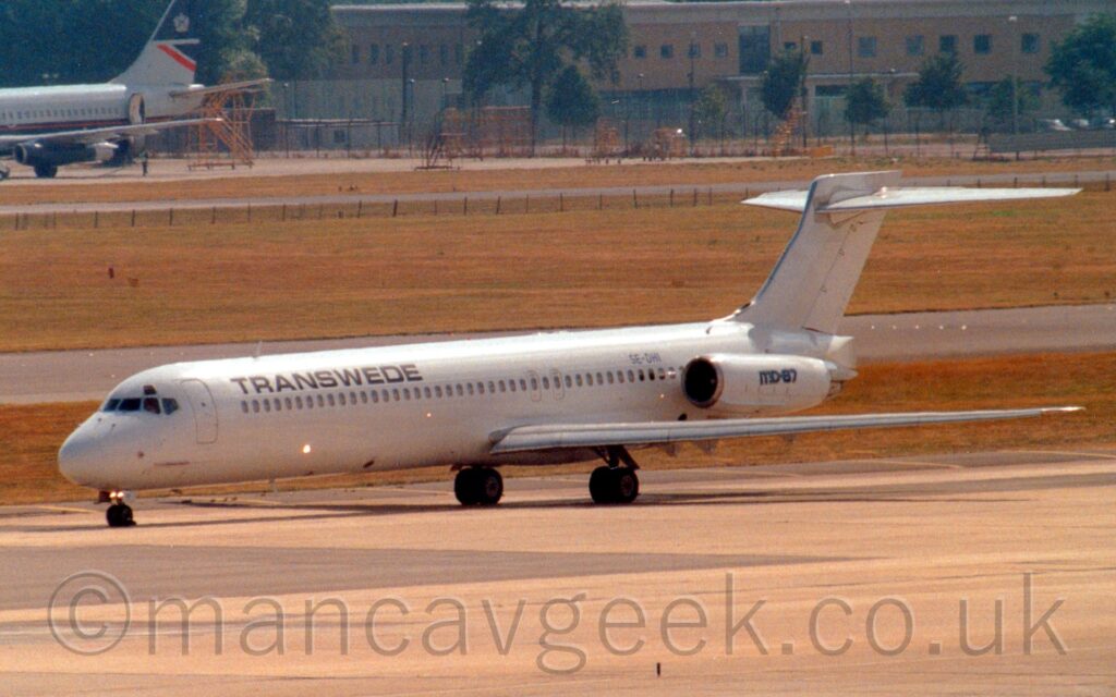 Side view of a twin engined jet airliner with the engines mounted on the sides of the rear fuselage, taxiing from right to left along a grey taxiway. The plane is almost entirely white, with only "TranSwede" titles on the upper forward fuselage, the black registration "SE-DHI" on the upper rear, and a black "MD-87" logo on the engine pods marring its blandness. Grey concrete apron fills the foreground, with a patch of grass in the background separating the plane from the runway, with more grass beyond that leading up to a large brown building in the distance on the right, and the rear fuselage and tail of a grey and dark blue airliner on the left.