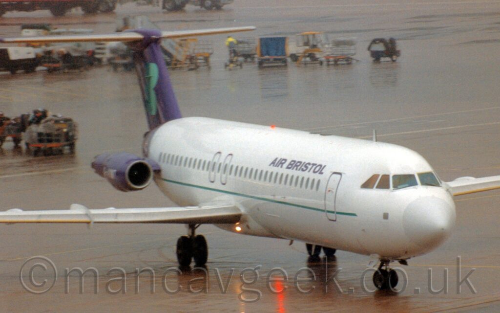 Front view of a twin engined jet airliner with the engines mounted on the sides of the rear fuselage, taxiing from left to right on a rather damp section of airport apron. The plane is mostly white, with purple tail and engine pods mounted on the sides of the rear fuselage. A thin green stripe runs along the lower body, under the passenger cabin windows, with purple "Air Bristol" titles on the upper forward fuselage, and the white registration "G-AVMT" on the sides of the engines. A bright landing light in the wing root and the red beacon in the centre of the underside reflect off the sodden apron surface in the foreground. More apron fills the rest of the frame, with lines of baggage carts stretching across he frame from the left in the background, and a grey and pink catering truck and white fuel truck in the top left corner.