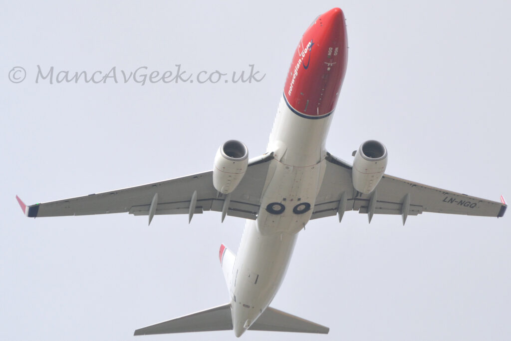 View from below of a twin engined jet airliner flying almost directly overhead at a very low altitude, with undercarriage tucked away, flaps extended from the rear of the wing, and the nose raised sharply, suggesting it has just taken off. The plane is mostly white, with a red nose and forward fuselage, and a thin blue band around the body directly aft of that. The white web address "Norwegian Dot Com" is on the lower forward fuselage. The black registration "LN-NGQ" is under the outer panels of the left wing. The tip of the tail and the up-turned wing-tips are red. Cloudy grey sky fills the rest of the frame.