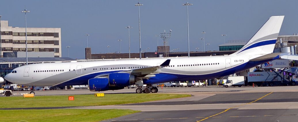 Side view of a white, 4 engined jet airliner taxiing from right to left along a grey taxiway. The plane has a grey and dark blue stripes running along the body, swooping up over the wing and back down again, and a smaller, similar stripe on the white tail. The engine pods under the wings are s deep blue, almost purple. The black registration "CS-TFX" is on the upper rear fuselage. Green grass fills the foreground on the left, with a black taxiway with a yellow line down the middle on the right. Grey airport buildings fill the background, with tall lighting towers climbing into the blue sky.