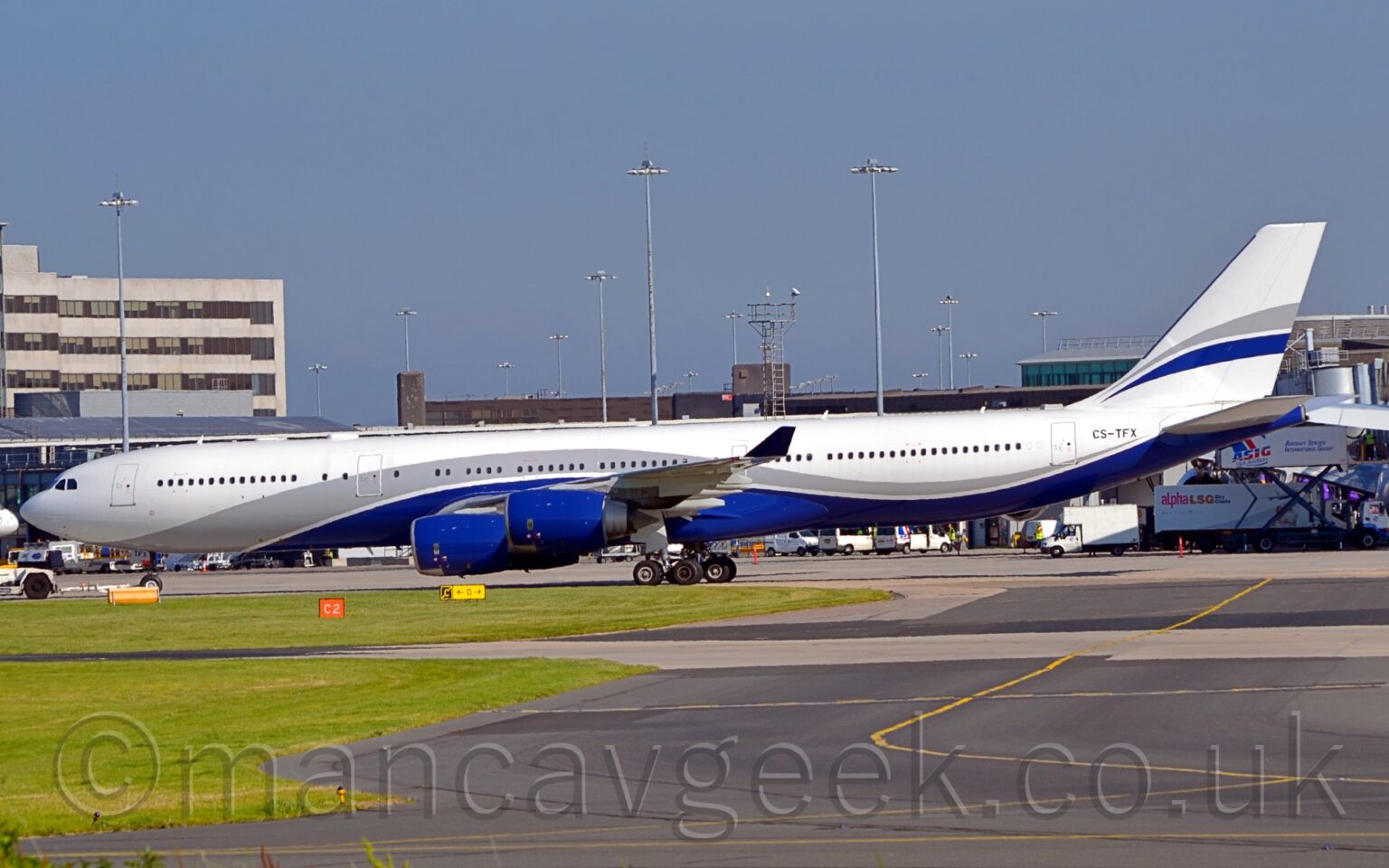 Side view of a white, 4 engined jet airliner taxiing from right to left along a grey taxiway. The plane has a grey and dark blue stripes running along the body, swooping up over the wing and back down again, and a smaller, similar stripe on the white tail. The engine pods under the wings are s deep blue, almost purple. The black registration "CS-TFX" is on the upper rear fuselage. Green grass fills the foreground on the left, with a black taxiway with a yellow line down the middle on the right. Grey airport buildings fill the background, with tall lighting towers climbing into the blue sky.