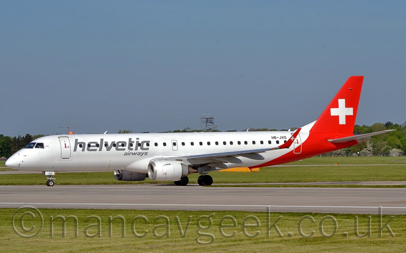 Side view of a twin engined jet airliner taxiing from right to left onto a grey runway. The plane is largely white, with a red rear fuselage and tail. There are grey "Helvetic" titles on the side of the forward fuselage, with smaller grey text "Airways" below. The black registration "HB-JVO" is on the upper rear fuselage, just forward of the rear cabin door. There is a large white cross on the red tail, as well as on the red up-turned wingtips. Green grass lines the runway in the foreground, with more grass in the background leading up to trees in the distance, under grey-blue sky.