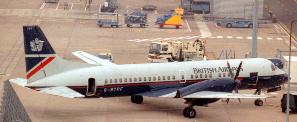 Elevated side view of a twin propellor-engined airliner parked facing to the left. The plane has a grey upper half, with black "British Airways" titles on the upper forward fuselage, and a dark blue ower half, with a red pin-stripe running along the body, ending in a half arrowhead at the front. The grey registration "G-BTPF" is on the lower rear fuselage, next to the open rear passenger door. The tail is dark blue on the top half, with a grey heraldic crest, and grey at the bottom, with a dark blue and red triangle towards the rear. Grey concrete apron fills the foreground, with a grey metal mesh blast barrier on the bottom left corner, while the background has more apron leading up to a terminal building whit a white airbridge with the Manchester Airport logo hanging off the side. The centre fuselage and engines of a white jet airliner can be seen in the top left corner. A re-fueling truck and a couple of laden baggage trucks can be seen parked in the background.
