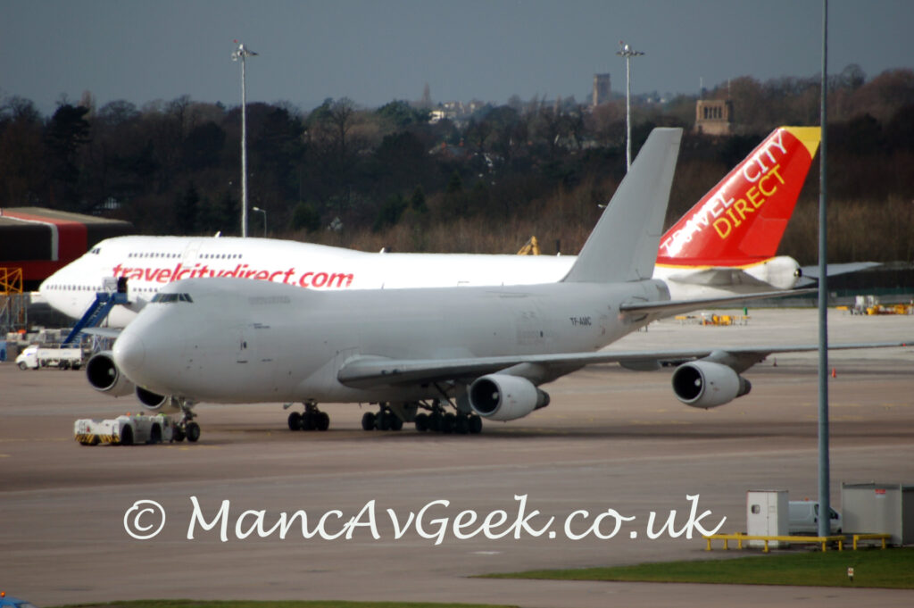 Side view of a very large, 4 engined jet airliner facing to the left but being pushed back to the right by a low-slung white truck attached to the nose-wheel. The plane is almost entirely white, with only the black registration "TF-AMC" on the rear fuselage breaking up the lack of colour. Grey concrete apron fills the foreground, with a green patch of grass in the lower right corner, and a grey lighting pole reaching up and out of the top of the frame. In the background, another white 4 engined jet airliner with a red and yellow tail and red web address "TravelCityDirect.Com" on the forward fuselage sits in bright sunlight, contrasting with this one which is in it's own shadow. Trees stretch across the background and into the distance, with buildings right at the back meeting grey-blue sky.