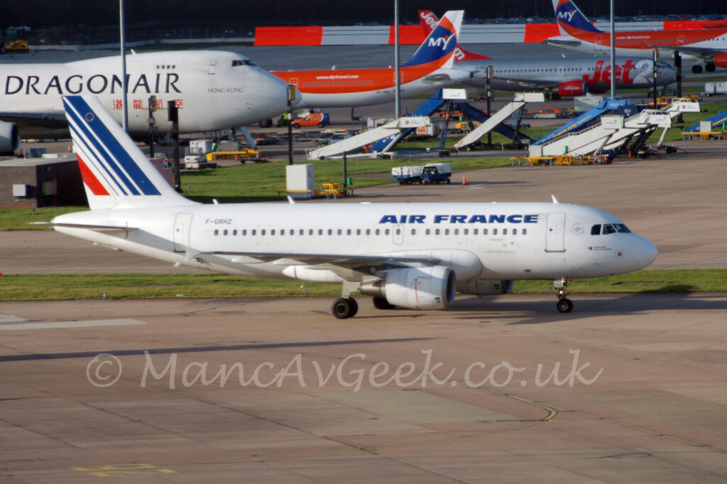 Side view of a twin engined jet airliner taxiing from left to right along a grey concrete taxiway. The plane is largely white, with blue "Air France" titles on the upper forward fuselage, and the black registration "F-GRHZ" on the upper rear, as well as the black letters "HZ" on the open nose wheel doors. A series of diagonal blue stripes are on the tail, getting thinner as they move to the rear, with a single red stripe right at the back. Grey concrete apron fills the foreground, with a strip of grass separating the plane from more apron in the background, with several other planes in various shades of white, silver, red, and blue in the distance, and a red and white-banded hoarding right at the back of the frame.