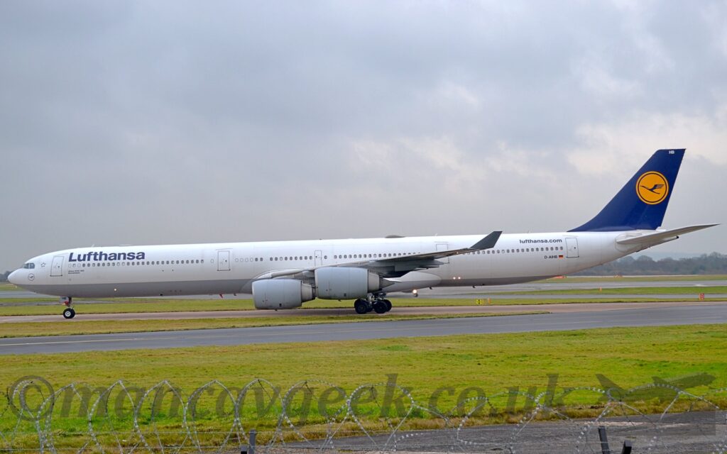 Side view of along and thin, 4 engined jet airliner taxiing from right to left along a grey taxiway. The plane is largely white, with a grey belly, and dark blue "Lufthansa" titles on the upper forward fuselage. The airlines web address "Lufthansa Dot Com" is on the upper rear fuselage, with the black registration "D-AIHB" below, next to a small German flag. The tail is dark blue, with a yellow circle containing a highly stylised image of a blue flying bird. The white letters "HB" are right at the top of the tail. Green grass fills the foreground, lining the side of the taxiway, with another taxiway cutting through in front, and razor wire on top of a fence right at the bottom of the frame. Fluffs of grey and white sky fill the rest of the frame.