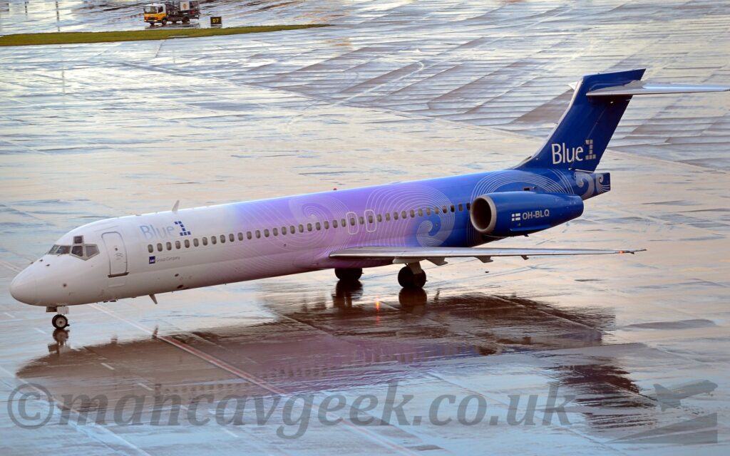 High side view of a twin engined jet airliner taxiing from right to left across a saturated section of airport apron. The plane is largely white, with a lilac section in the middle, over the wing root, and blue at the rear, both with swirly white piping, and grey and blue "Blue 1" titles on the upper forward fuselage. Smaller, white and grey "Blue1" titles are on the blue tail, and the white registration "OH-BLQ" is on the side of the engine pods, mounted on the sides of the rear fuselage, next to a small Finnish flag. The plane is reflected in puddles on the rather wet apron in the foreground, which suggests it has recently been raining, if it is not still doing so, with more wet apron filling the background.