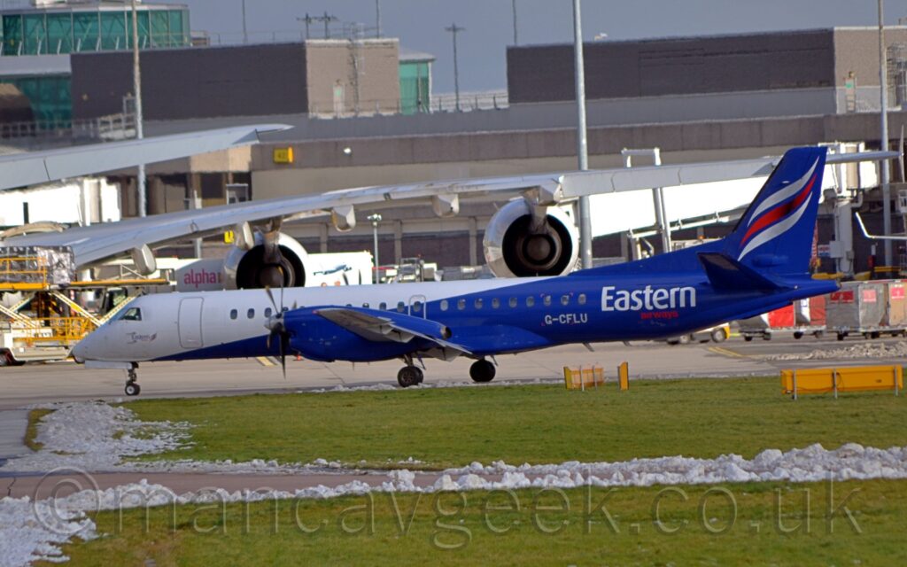 Side view of a white and blue, twin propellor-engined airliner taxiing from right to left. The front of the plane is all white, with the rear fuselage and tail all dark blue. There are white and red "Eastern Airways" titles on the rear fuselage, as well as the white registration "G-CFLU". There are white, red, white, diagonal stripes on the tail. Green grass fills the foreground, with clumps of snow lining the sides of a taxiway. A grey terminal building stretches across the frame, behind the long wing of a very large jet airliner with a pair of large engines hanging off the wing. Grey sky fills the rest of the frame.