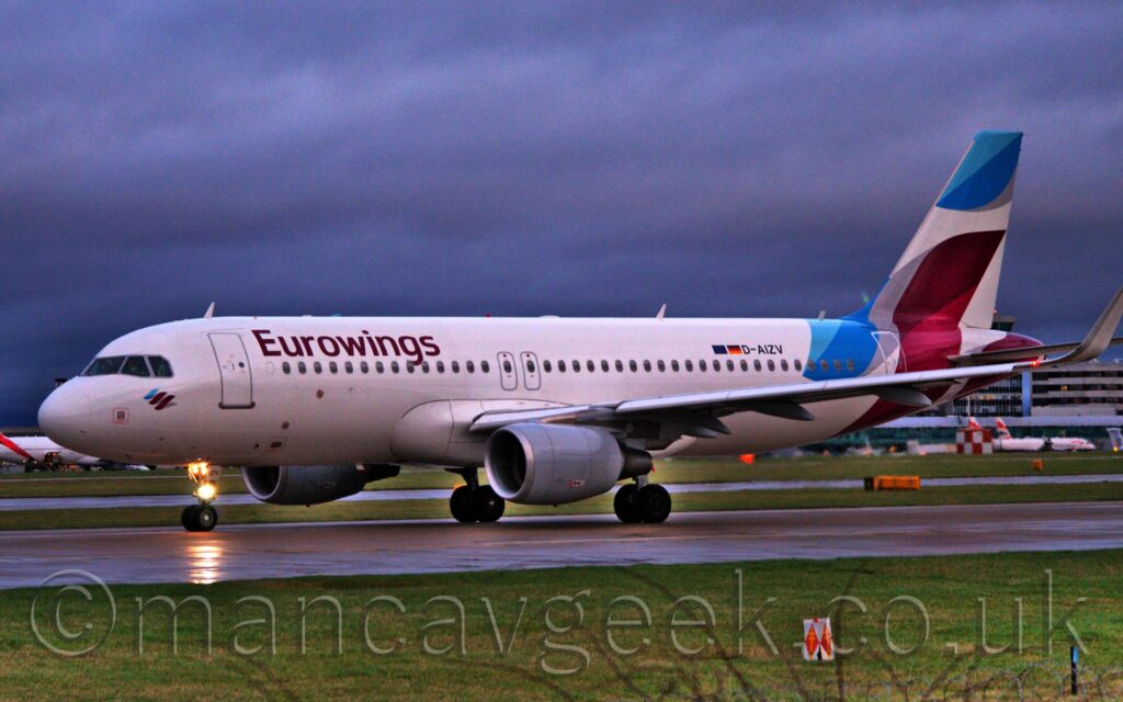 Side view of a twin engined jet airliner taxiing from right to left along a black runway. The plane is mostly white, with mauve "EuroWings" titles on the upper forward fuselage, and seemingly random light blue, grey and mauve stripes on the tail. The black registration "D-AIZV" is on the upper rear fuselage next to small German and EU flags. Bright lights shine forwards on the nosewheel gear leg. Green grass lines the sides of the runway in the foreground, with more grass in the background leading up to a large, grey terminal building in the distance, under dreary grey sky.