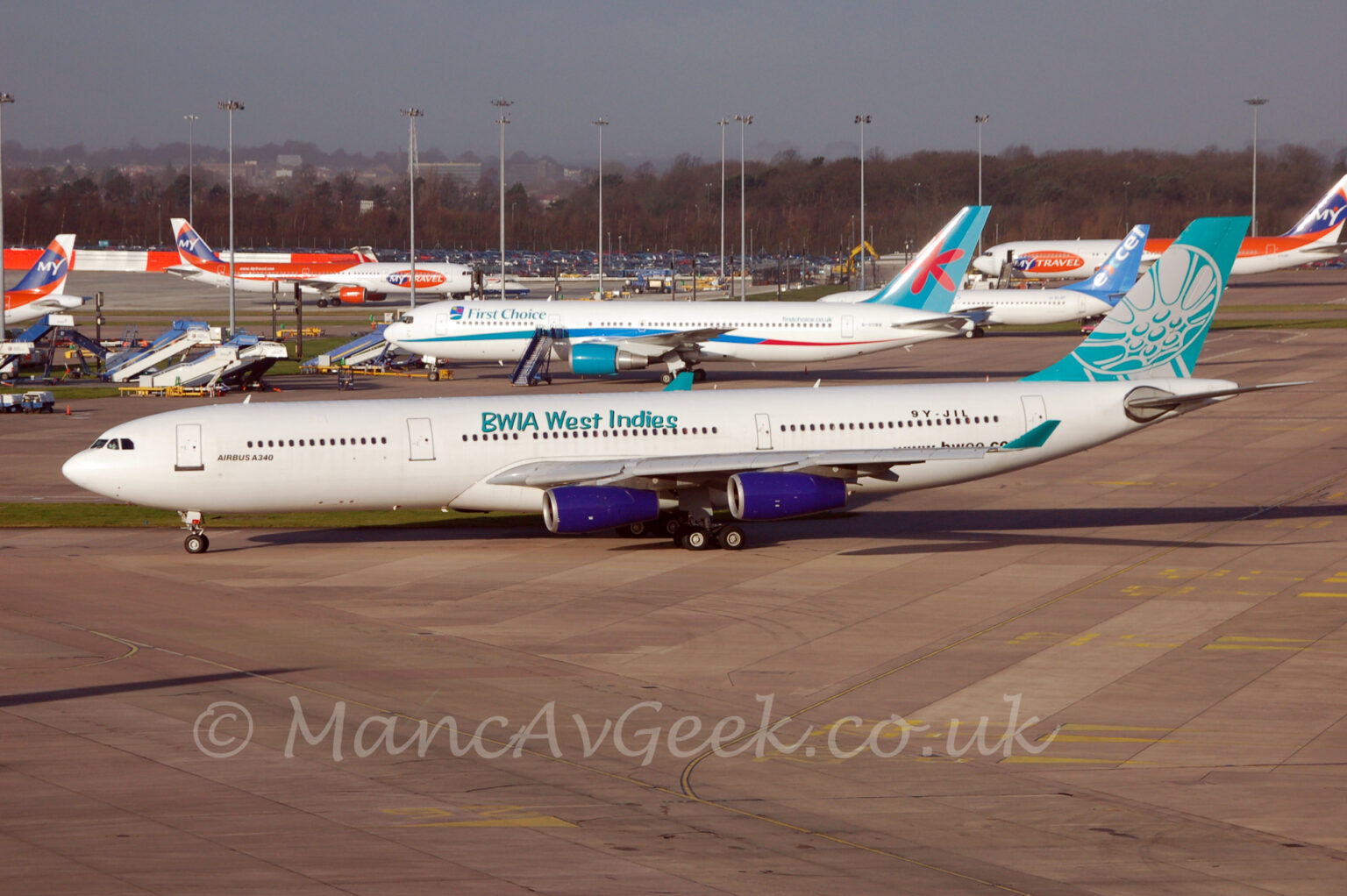 Side view of a 4-engined jet airliner taxiing from right to left at a busy airport. The plane is mostly white, with blue-green "BWIA West Indies" titles on the upper forward fuselage, over the wings, and black "Airbus A340" text on the lower forward fuselage, just aft of the forward cabin door. The black registration "9Y-JIL" is on the upper rear fuselage, with the web address "www.bwee.com" on the lower rear. The green-blue tail has a stylised, white steel drum filling most of the space. The engine pods, mounted under the wings, are da deep purple, while the up-turned wing-tips being a similar colour to the tail, possibly a bit greener. Grey concrete apron fills the foreground, with 5 jet airliners, some white and blue, others white and orange, parked on apron in the background, stuck amongst some tail lighting poles, with trees in the distance under flat grey sky.