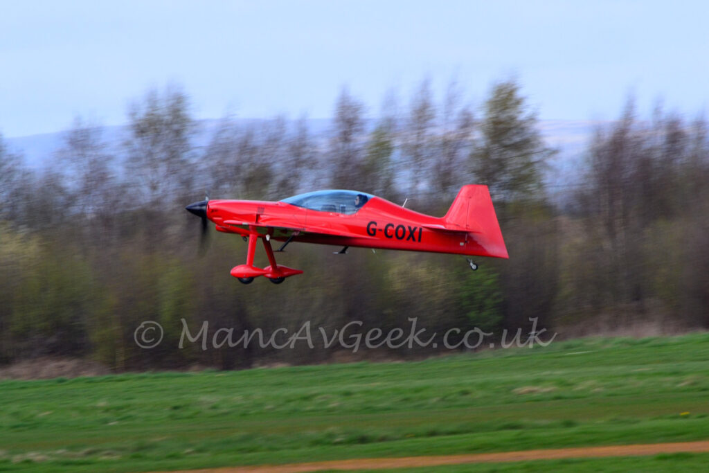 Side view of a small, sleek, single engined light aircraft flying from right to left t a very low altitude, just a few metres above a grass runway. The plane is a bright scarlet red, with a large glazed canopy over the 2-seat cockpit, with only the rear seat filled. Long, spindly undercarriage legs extend down from the front of the wings, with large red spats over the main wheels. The spinner on the propellor is a gloss black, , as is the registration "G-COXI" on the sides of the rear fuselage. Greengrass fills the foreground at the bottom of the frame, with trees filling the background, with rolling hills and pale blue sky in the distance.