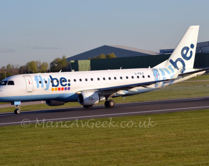 Side view of a twin engined jet airliner moving at speed from right to left along a black runway. The plane is mostly white, with a pale blue belly, and large light and dark blue "FlyBe" titles above 6 coloured dots (2 yellow, 2 red, 2 purple) on the forward fuselage, a design repeated on the sides of the white engine pods mounted under the wings with additional ".com" text at the end. The light and dark blue "FlyBe" titles are repeated in a larger form but without the dots on the rear fuselage and tail. The black registration "G-FBJI" is on the upper rear fuselage. Green grass fills the foreground, lining the sides of the runway. A pair of large grey hangars dominate the background on the right, behind a large green metal wall. Pale blue sky fills the rest of the frame.