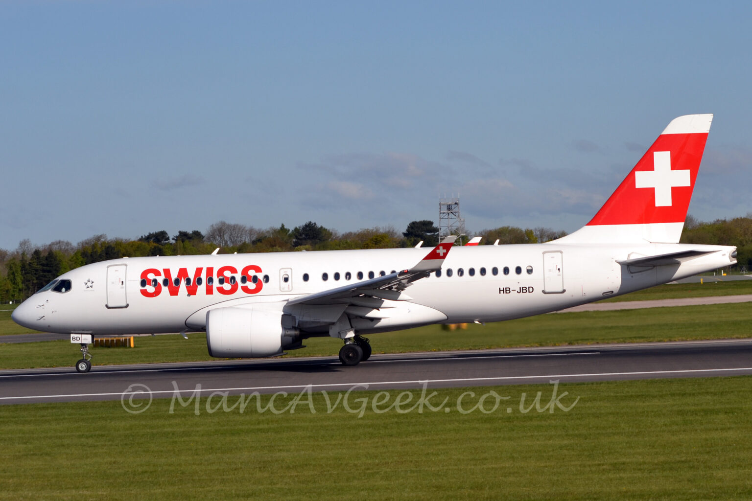 Side view of a white, twin engined jet airliner asccelerating from right to left as it starts it's take-off run on a black runway. The plane has large red "Swiss" titles on the forward fuselage, covering the forward passenger cabin windows, and the black registration "HB-JBD" on the lower rear fuselage. A thick red stripe on the white tail has a large white cross in the middle, a design replicated on the up-turned wingtips. Green grass fills the foreground, lining the sides of the black runway, with more grass in the background leading up to trees in the distance, under blue sky.