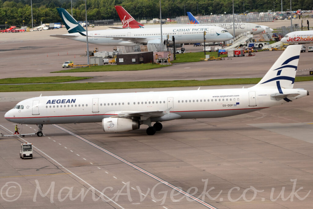 Side view of a white, twin engined jet airliner facing to the left but being pushed back to the right by an almost out-of-frame tug attached to the nose-wheel by a long white pole. The plane has a grey belly, and a thin red pinstripe running along the border between the white and grey. There are dark blue "Aegean" titles on the upper forward fuselage, and the black registration "SX-DGP" on the lower rear fuselage, next to small Greek and EU flags. The airline's web address is on the upper rear fuselage, just forward of the rear door, with small text "Airbus A321" just aft of the door. The outline of a dark blue circle is on the side of the engine pods, overlaid with a couple of stylised birds drawn as single lines, a logo that is partially repeated in a much larger form on the white tail, only the left part of the circle along with the birds wings being shown. Grey concrete apron fills the foreground, with a small, white, baggage truck parked on the left of the frame, and a man in red trousers and a yellow hi-viz vest walking alongside the plane just above the tug. A strip of green grass separates this plane from more apron in the background, with tall lighting poles reaching out of frame, and several mostly white jet airliners parked, with trees in the distance.