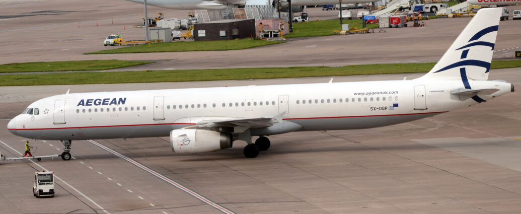 Side view of a white, twin engined jet airliner facing to the left but being pushed back to the right by an almost out-of-frame tug attached to the nose-wheel by a long white pole. The plane has a grey belly, and a thin red pinstripe running along the border between the white and grey. There are dark blue "Aegean" titles on the upper forward fuselage, and the black registration "SX-DGP" on the lower rear fuselage, next to small Greek and EU flags. The airline's web address is on the upper rear fuselage, just forward of the rear door, with small text "Airbus A321" just aft of the door. The outline of a dark blue circle is on the side of the engine pods, overlaid with a couple of stylised birds drawn as single lines, a logo that is partially repeated in a much larger form on the white tail, only the left part of the circle along with the birds wings being shown. Grey concrete apron fills the foreground, with a small, white, baggage truck parked on the left of the frame, and a man in red trousers and a yellow hi-viz vest walking alongside the plane just above the tug. A strip of green grass separates this plane from more apron in the background, with tall lighting poles reaching out of frame, and several mostly white jet airliners parked, with trees in the distance.