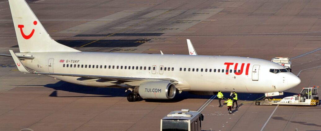 Side view of a twin engined jet airliner facing to the right but being pushed back to the left by a low-slung white tug which has lifted it's nose-wheel off the ground. The plane is predominantly white, with red "TUI" titles on the upper forward fuselage, and the black registration "G-TUKF" next to a small UK flag on the upper rear. The white tail has a red logo that looks like a smushed-up winky-face made up of the letters "T", "U", and "I", with a smaller version of this logo appearing on the up-turned wing-tips. The engine pods mounted under the wings are grey, with the black web address "TUI Dot Com" on the side. Grey concrete apron fills the foreground, with 3 p3opl3 in hi-viz jackets walking alongside the plane as it moves backwards, and a silvery-coloured airport bus waiting on a roadway marked out with white lines on the surface. More apron stretches into the distance, with a load of vehicles parked in front of a terminal building in the distance.