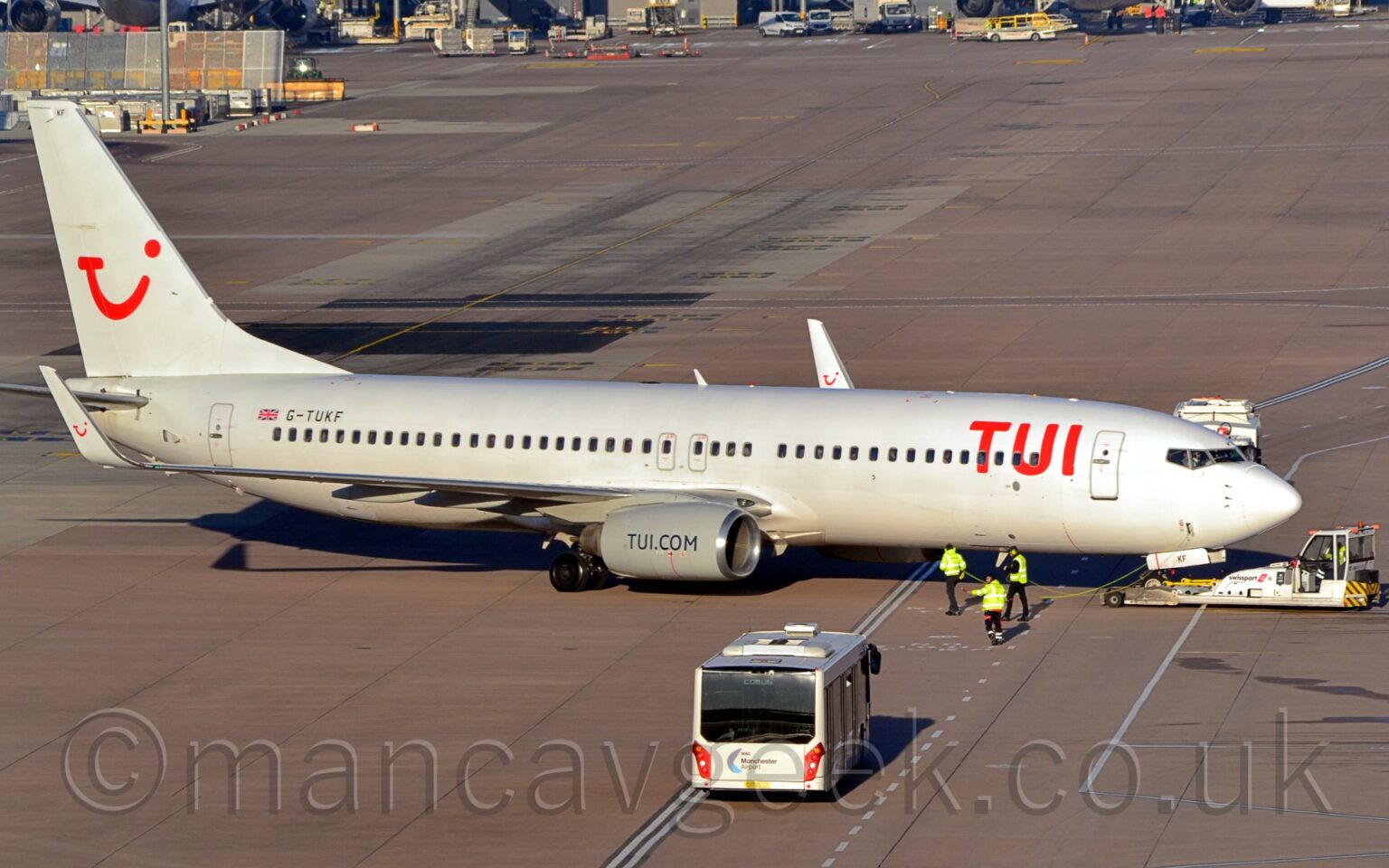 Side view of a twin engined jet airliner facing to the right but being pushed back to the left by a low-slung white tug which has lifted it's nose-wheel off the ground. The plane is predominantly white, with red "TUI" titles on the upper forward fuselage, and the black registration "G-TUKF" next to a small UK flag on the upper rear. The white tail has a red logo that looks like a smushed-up winky-face made up of the letters "T", "U", and "I", with a smaller version of this logo appearing on the up-turned wing-tips. The engine pods mounted under the wings are grey, with the black web address "TUI Dot Com" on the side. Grey concrete apron fills the foreground, with 3 p3opl3 in hi-viz jackets walking alongside the plane as it moves backwards, and a silvery-coloured airport bus waiting on a roadway marked out with white lines on the surface. More apron stretches into the distance, with a load of vehicles parked in front of a terminal building in the distance.