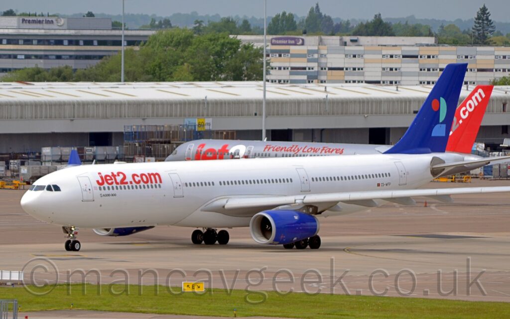 Side view of a twin engined jet airliner taxiing from right to left along a grey concrete taxiway. The plane is mostly white, with red "Jet2.com" titles on the upper forward fuselage, and much smaller black text "Airbus A330-300" under the forward passenger windows. The tail is a deep blue, with a heavily stylised number "2" in red, light blue, and aquamarine. The engine pods under the wings are the same deep blue. In the background, another jet airliner, this one grey with red and white "Jet2" titles and red text "Friendly low fares" on the upper fuselage sits in the middle of the frame, in front of a wide, grey building that stretches the width of the frame. A pair of larger grey buildings are in the distance, both with "Premier Inn" signs near the top. Grey sky fills what little space is left at the top.