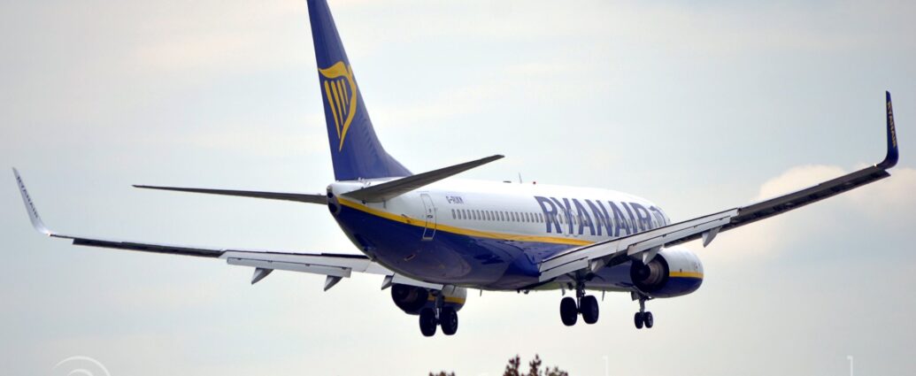 Side view of a twin engined jet airliner flying from left to right at a very low altitude, with undercarriage lowered and flaps drooping down from the rear of the wings, suggesting it is about top land. The plane is mostly white, with a dark blue belly, and a yellow stripe running along the body below the passenger cabin windows. There are large, dark blue "RyanAir titles on the sides of the forward fuselage, and the registration "G-RUKM" on the upper rear. The tail is dark blue, with a large, yellow, winged Irish harp in the middle. The engine pods are similar to the body, white on top, blue on the bottom, and a yellow stripe through the middle. The tops of trees can be seen at the bottom of the frame, with cloudy sky with patches of pale blue filling the rest.