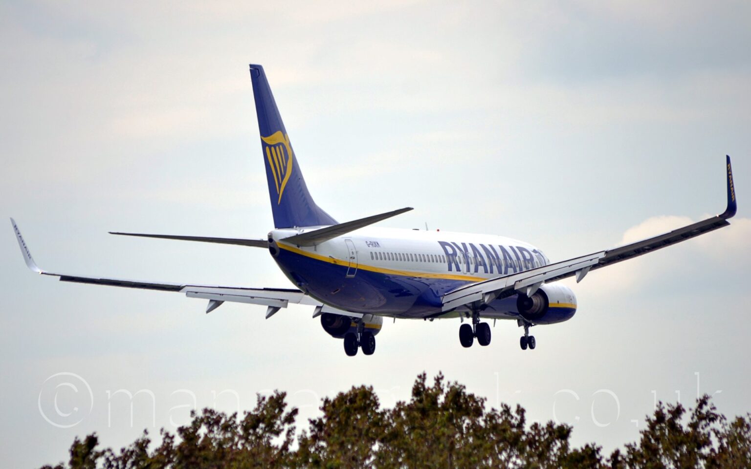 Side view of a twin engined jet airliner flying from left to right at a very low altitude, with undercarriage lowered and flaps drooping down from the rear of the wings, suggesting it is about top land. The plane is mostly white, with a dark blue belly, and a yellow stripe running along the body below the passenger cabin windows. There are large, dark blue "RyanAir titles on the sides of the forward fuselage, and the registration "G-RUKM" on the upper rear. The tail is dark blue, with a large, yellow, winged Irish harp in the middle. The engine pods are similar to the body, white on top, blue on the bottom, and a yellow stripe through the middle. The tops of trees can be seen at the bottom of the frame, with cloudy sky with patches of pale blue filling the rest.