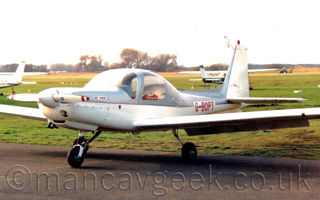 Side view of a single engined light aircraft parked facing to the left. The planes upper surfaces are a metallic, silvery-grey, with white lower surfaces, and red piping. The registration "G-BOPT" is on the rear fuselage in red. The tail is white, with a grey leading edge, and a red beacon on the top. A large grass airfield fills the background, leading up to trees in the distance, with a couple of other light aircraft parked on the grass. Bright but hazy sky fills the rest of the frame.