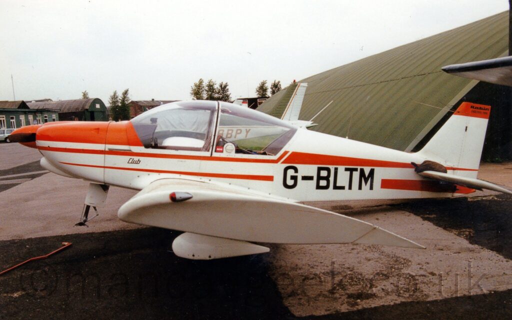 Side view of a single-engined light aircraft parked facing to the left, with it's tail weighed down so it's nose, minus the nose-wheel, is raised in the air. The plane is mostly white, with bright orange stripes running along the body, and the black registration "G-BLTM" on the lower rear fuselage. There is a green hangar in the background on the right, with more green buildings in the distance on the right, under flat grey skies.