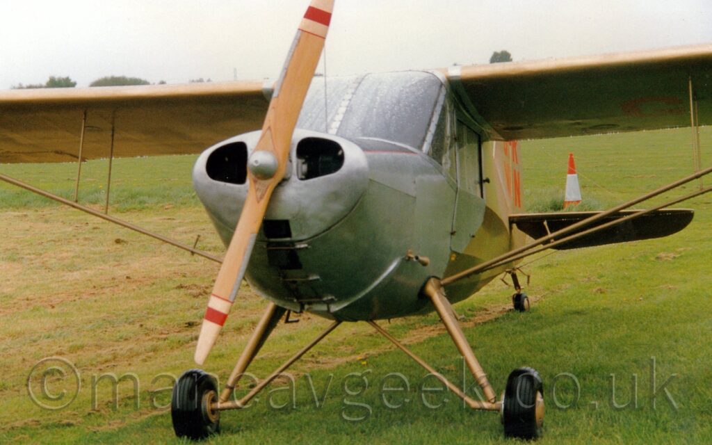 Almost head-on view of a single-engined, high-winged light aircraft parked facing to the left of the camera on a grassy field. The plane is mostly in a golden finish on the body and wings, with silver engine cowlings and doors. The wooden propellor has white, red, and white bands on the tips. The red registration "G-RWIN" is visible on the tail. Green grass fills the foreground and most of the background, with trees in the distance visible over the tops of the wings, meeting grey sky.